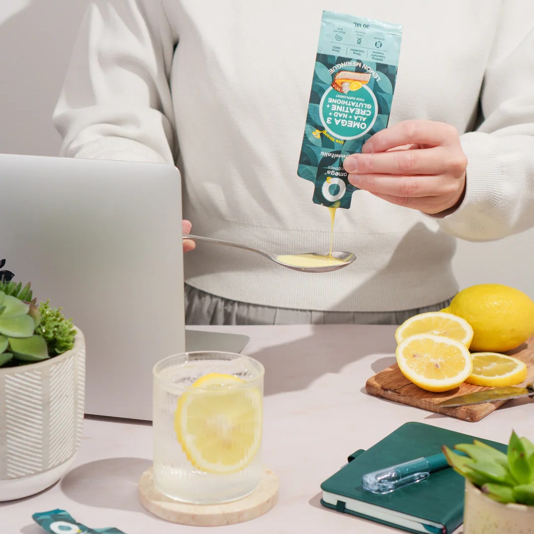 Person pouring lemon-flavored omega supplement from a sachet onto a spoon beside a laptop, glass of lemon water and fresh lemons.